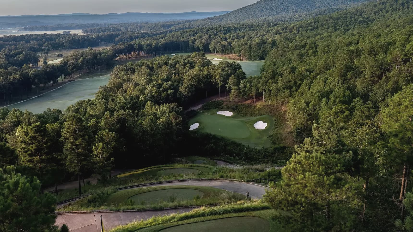 Aerial view of a green and bunker complex at FarmLinks at Pursell Farms, framed by Alabama hardwood forest and a distant ridgeline.