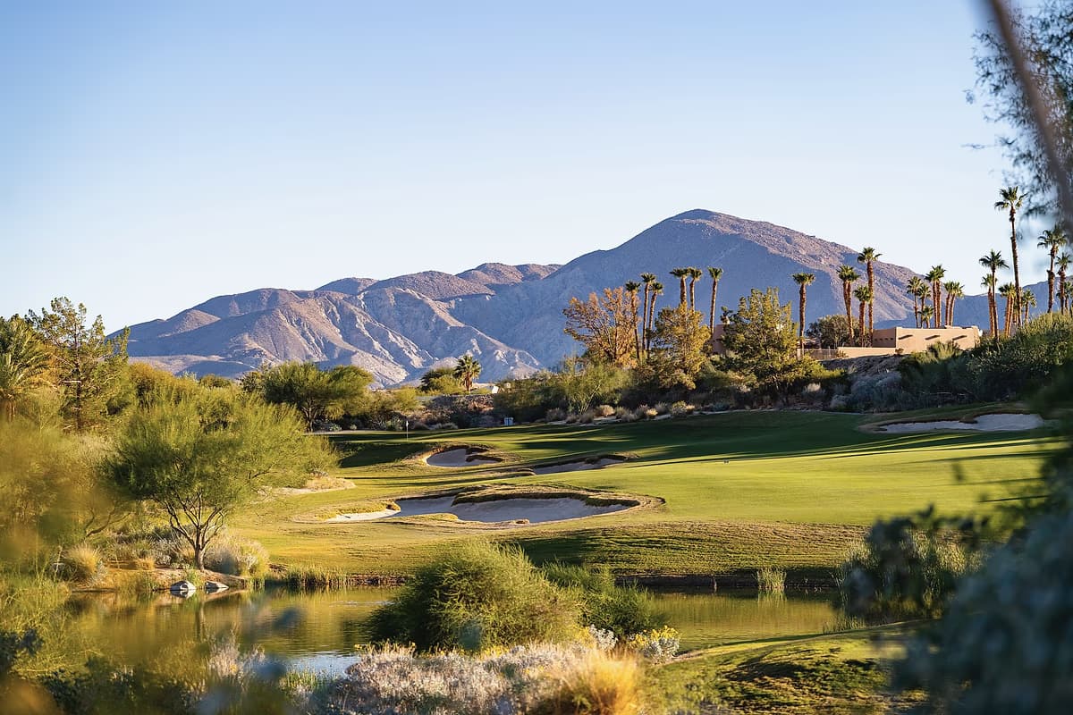 A desert mountain rising behind palms in golden light at Rams Hill Golf Club, with water and bunkers in the foreground.